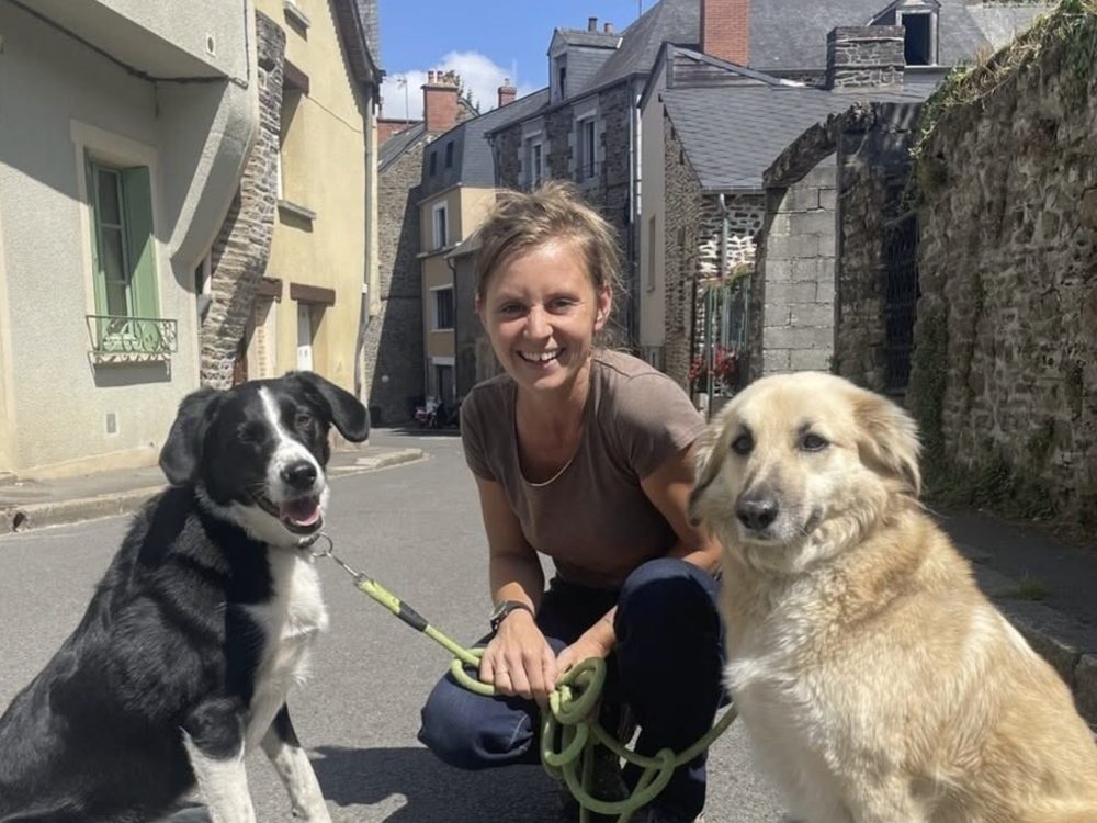 Amélie Owieczka souriante, accroupie dans une rue de Fougères entre deux chiens